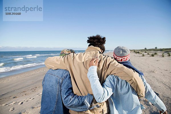Drei Freunde gehen am Strand entlang  Arme umeinander  Rückansicht