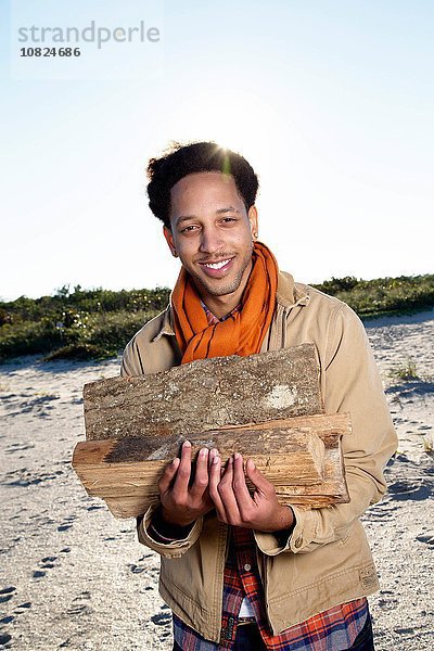 Porträt eines jungen Mannes am Strand  der Feuerholz hält  lächelnd