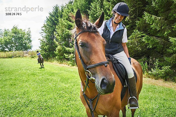 Reife Frau auf dem Rücken eines Pferdes mit Reitkappe und Stiefeln beim Anblick eines lächelnden Pferdes.