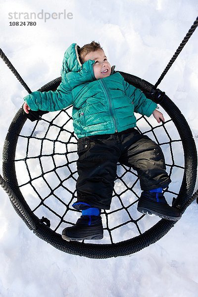 Overhead-Ansicht des Vorschulkindes auf Hängemattenschaukel im Schnee liegend mit lächelndem Blick