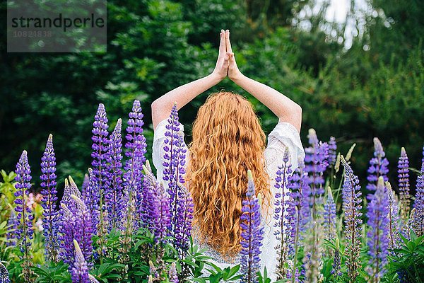 Rückansicht einer jungen Frau  die mit den Händen zusammen inmitten von lila Wildblumen meditiert.