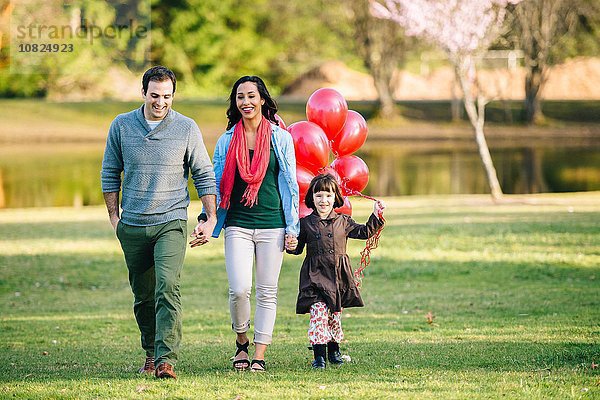 Junges Paar und Tochter mit einem Haufen roter Luftballons beim Spaziergang im Park