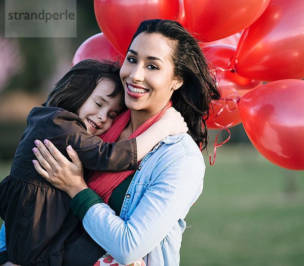 Porträt eines Mädchens mit einem Haufen roter Luftballons  die die Mutter im Park umarmen.