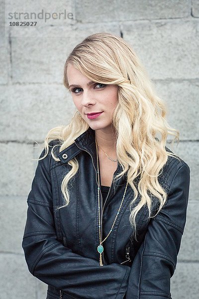 Junge blondhaarige Frau vor der Breeze-Blockwand in Lederjacke mit Blick auf die Kamera lächelnd