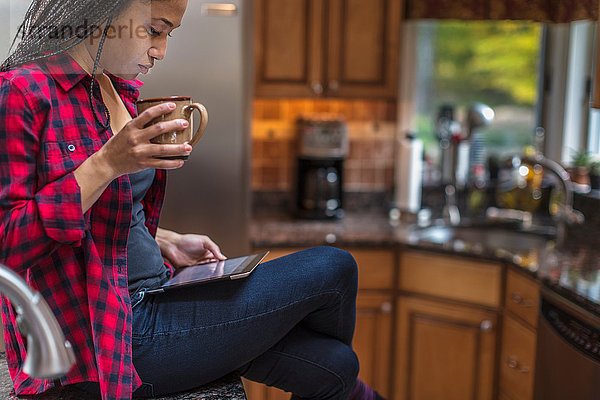 Mittlere erwachsene Frau  die in der Küche sitzt und ein digitales Tablett benutzt.