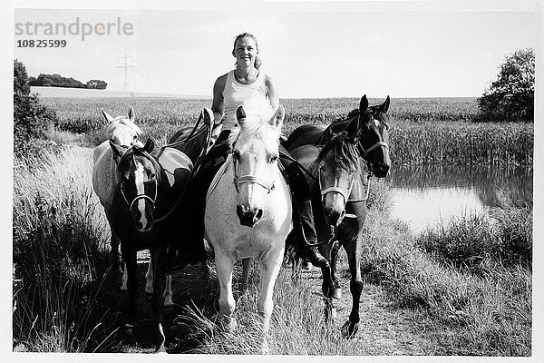 Schwarzweiß-Bild von einer Frau  die sechs Pferde auf einem Feldweg reitet und führt.