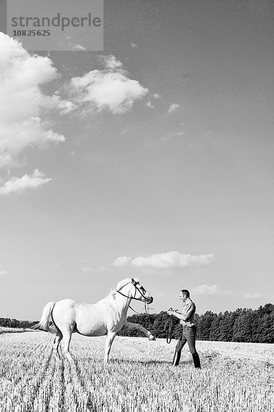 Schwarzweißbild eines Mannes  der weißes Pferd im Feld trainiert.