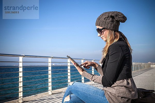 Seitenansicht der jungen Frau am Meer sitzend mit Hilfe eines digitalen Tabletts