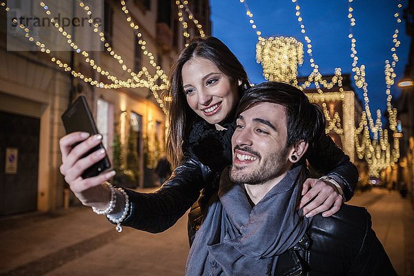 Junger Mann gibt junge Frau Huckepack mit Smartphone zu nehmen Selfie  lächelnd