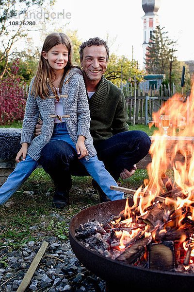 Mädchen sitzt auf Vaters Schoß im Garten und beobachtet die Feuerstelle.