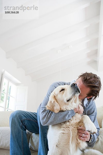 Junger Mann auf dem Wohnzimmersofa umarmt Golden Retriever Dog