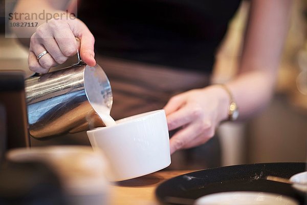 Abgeschnittener Schuss einer Kellnerin  die Milch in eine Kaffeetasse im Café gießt.