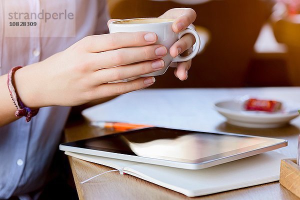Hände einer jungen Frau  die eine Kaffeetasse mit einem digitalen Tablett hält.