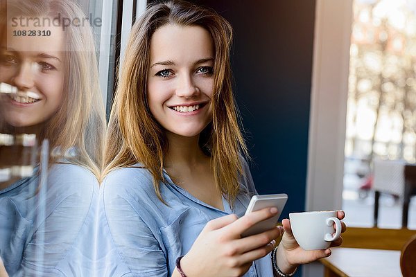 Junge Frau hält Kaffeetasse und Smartphone und schaut lächelnd in die Kamera