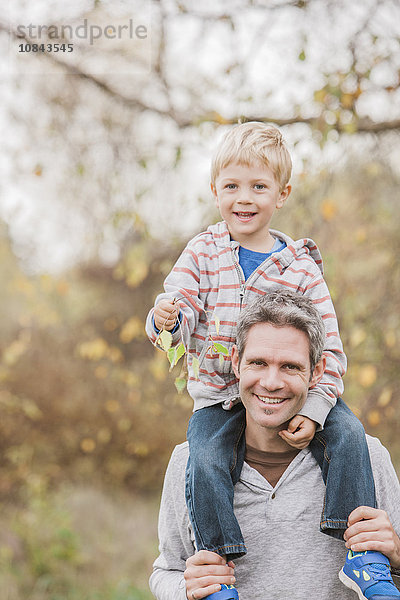 Portrait lächelnder Vater mit Kleinkind-Sohn auf den Schultern im Herbstpark