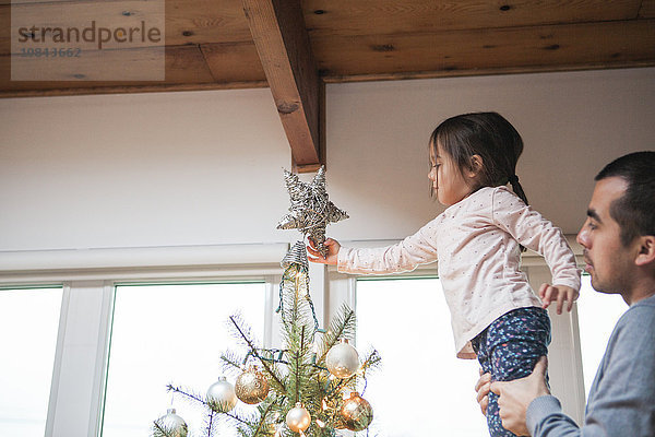 Vater hebt Kleinkind Mädchen setzt Stern auf Weihnachtsbaum