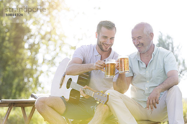 Vater und erwachsener Sohn rösten Bierkrüge und spielen Gitarre im Freien.