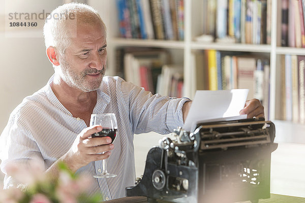 Senior Mann trinkt Wein an der Schreibmaschine im Arbeitszimmer