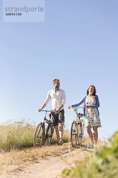 Junges Paar geht mit dem Fahrrad auf dem Feldweg unter dem sonnigen blauen Himmel spazieren