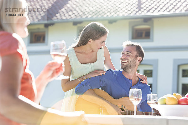 Lächelndes junges Paar spielt Gitarre und trinkt Wein auf der Sonnenterrasse.