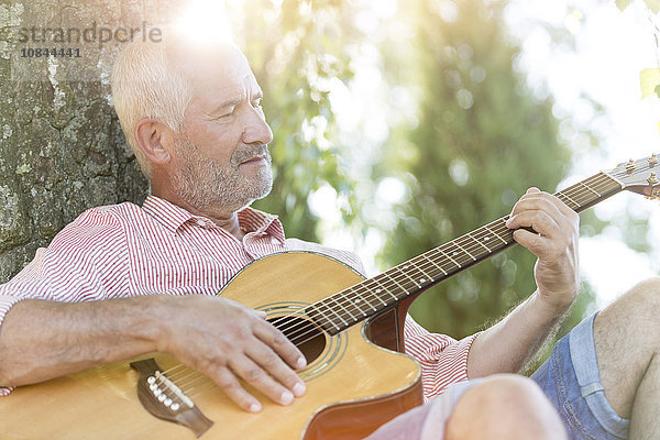 Senior Mann spielt Gitarre gegen Baumstamm