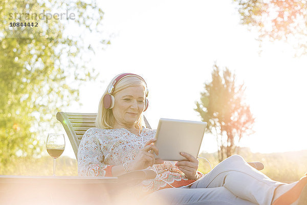 Seniorin mit Kopfhörer und Wein mit digitalem Tablett auf Sessel im sonnigen Hinterhof