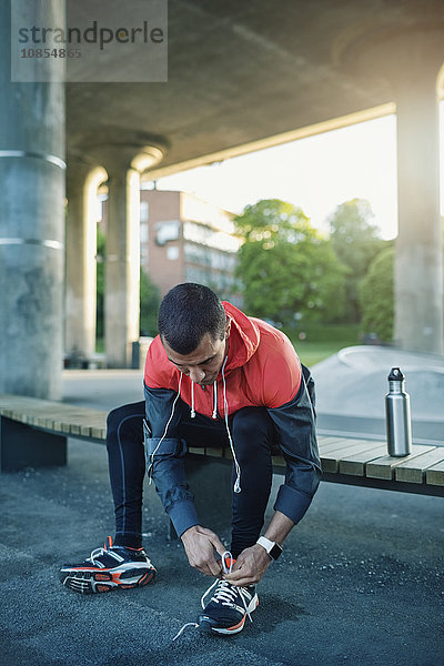 Man tying shoelace while sitting on bench under bridge