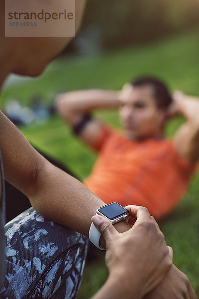 Woman touching smart watch while man doing sit ups at park