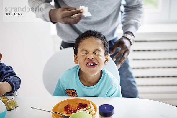 Boy making face while having food at table with father standing in background