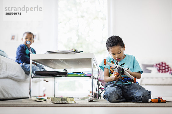 Boy examining model airplane at home with brother sitting on sofa in background