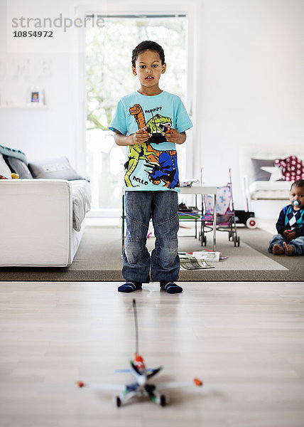 Boy playing with remote controlled airplane at home with brother sitting in background