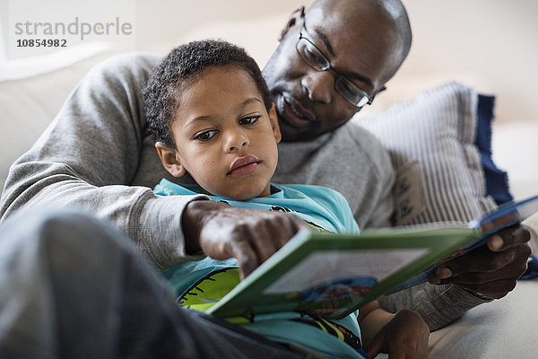 Father assisting bored son in reading book on sofa at home