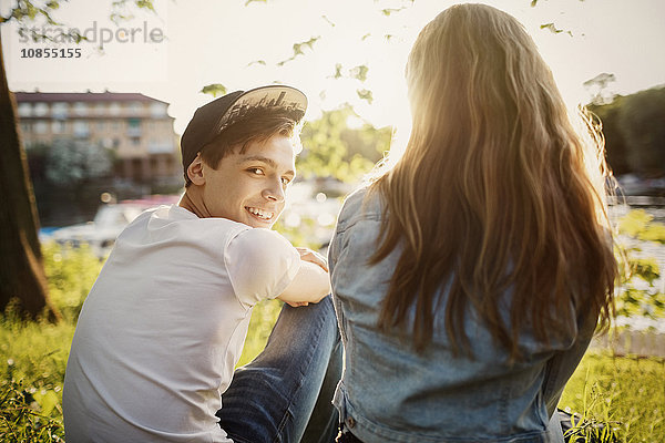 Rear view portrait of happy teenager sitting with female friend at lakeshore