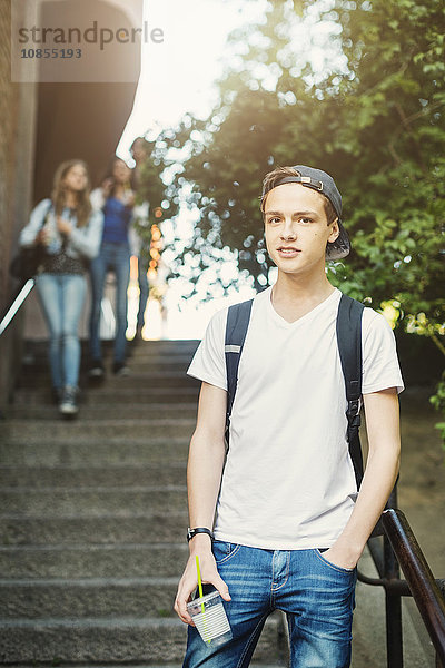 Portrait of confident teenager holding disposable glass while standing on steps with friends in background