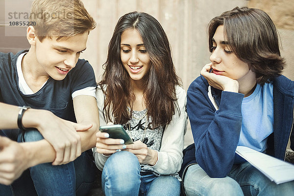 Happy girl using smart phone while sitting with male friends on steps