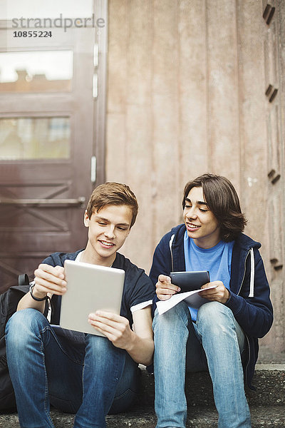 Happy male teenagers using digital tablet while sitting on steps outdoors