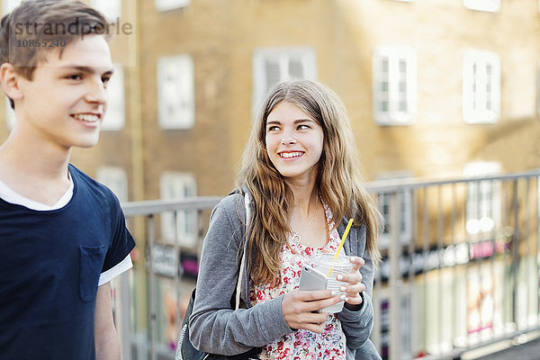 Happy teenager holding disposable glass and smart phone while looking at friend