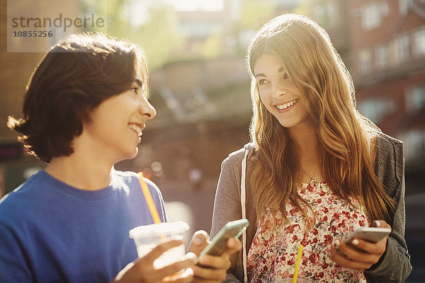 Happy teenagers looking at each other while holding smart phones outdoors