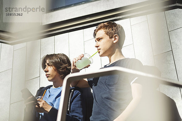 Boy looking away while drinking juice with friend holding smart phone outdoors