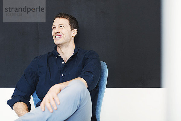 Happy mid adult businessman sitting on chair against wall in office