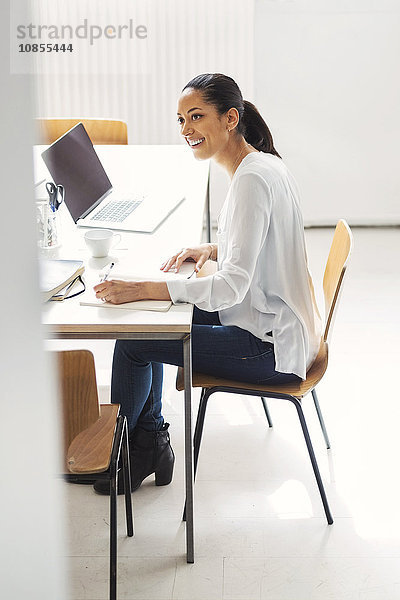 Side view of happy young businesswoman writing in book at desk in office