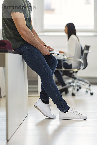 Low section of young businessman sitting on desk with colleague working in background at office