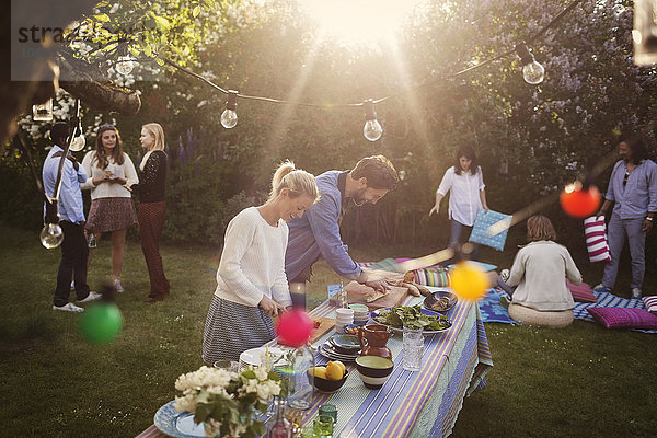 Man and woman preparing food at dining table with friends in background during summer party