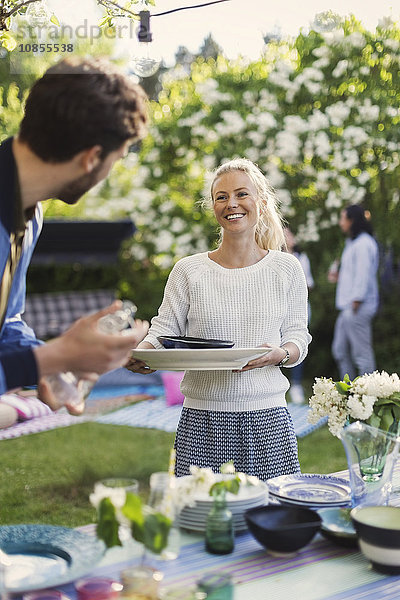 Happy young woman holding plates while looking at friend in yard during summer party