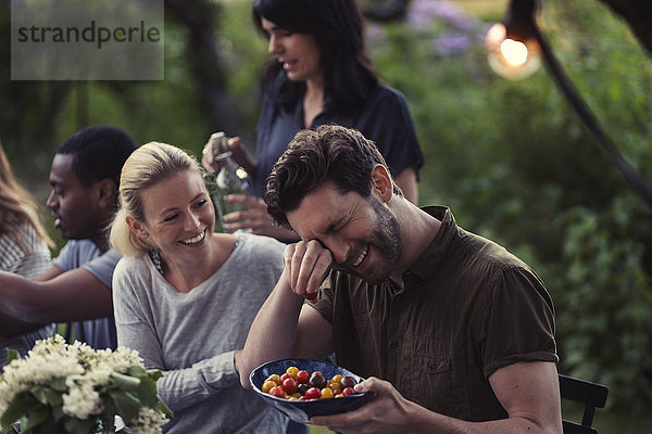 Happy woman looking at man rubbing eyes while holding tomato bowl at dinner party in yard