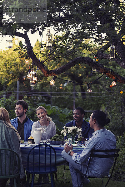 Happy multi-ethnic friends sitting at outdoor food table during evening party