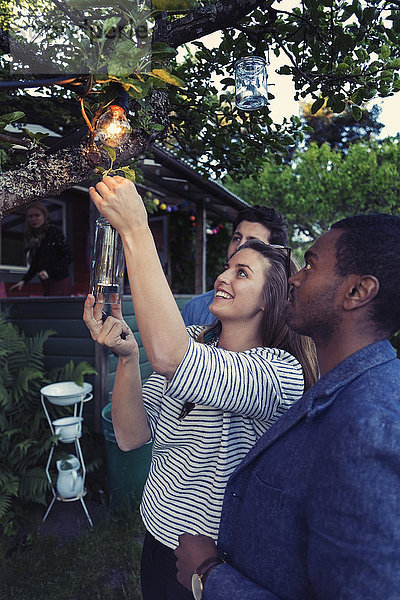 Happy woman hanging candle bottle on branch at yard for party