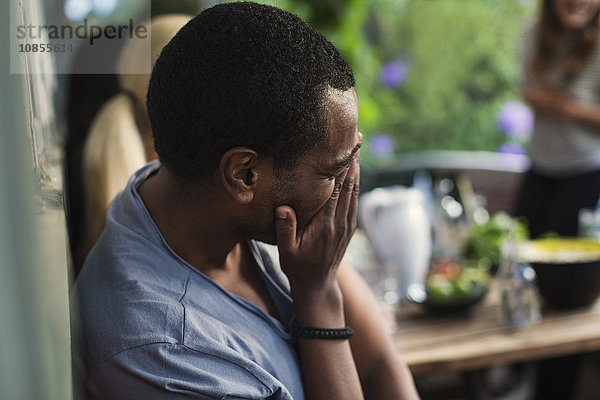 Playful man covering face while sitting at porch in summer house