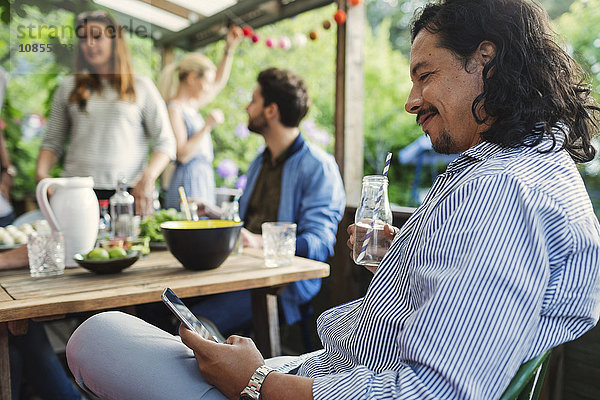 Side view of smiling man using smart phone during summer party in log cabin