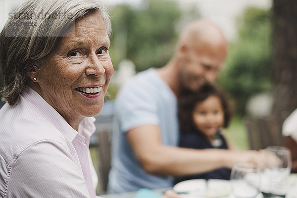 Side view portrait of happy senior woman with family having food at outdoor table in background